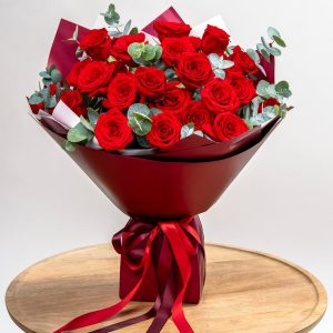 Buchet de trandafiri roșii cu eucalipt, aranjat în ambalaj roșu elegant cu panglici satin.